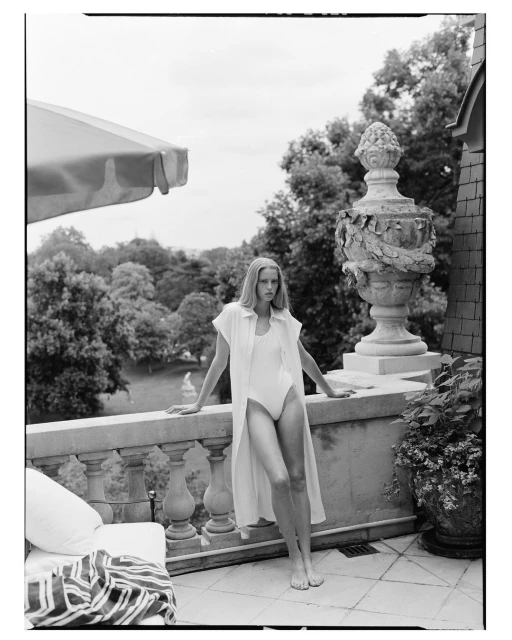 Monochrome shot of an adult woman in a light-colored swimsuit and cover-up standing on a stone patio with a garden and cloudy sky background.