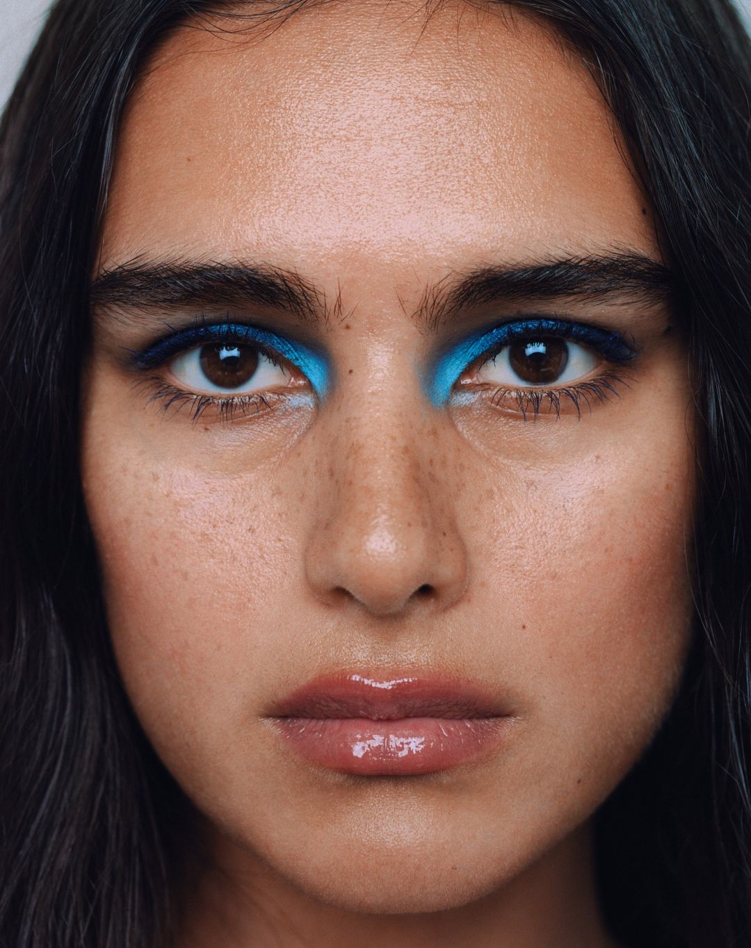 Close-up portrait of a woman with brown hair and eyes, wearing bright blue eye shadow and glossy lip gloss, showcasing a beauty shot.
