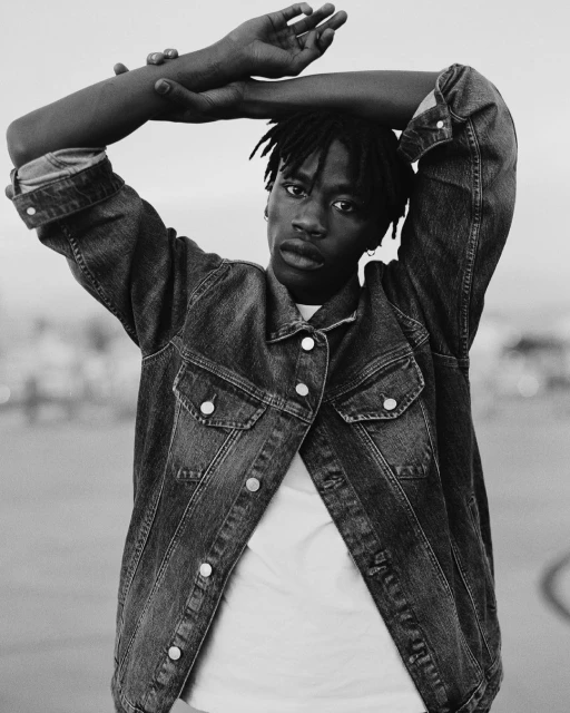 Monochrome portrait of an African American adult male with dreadlocks wearing a jean jacket, arms raised over head, outdoor setting.