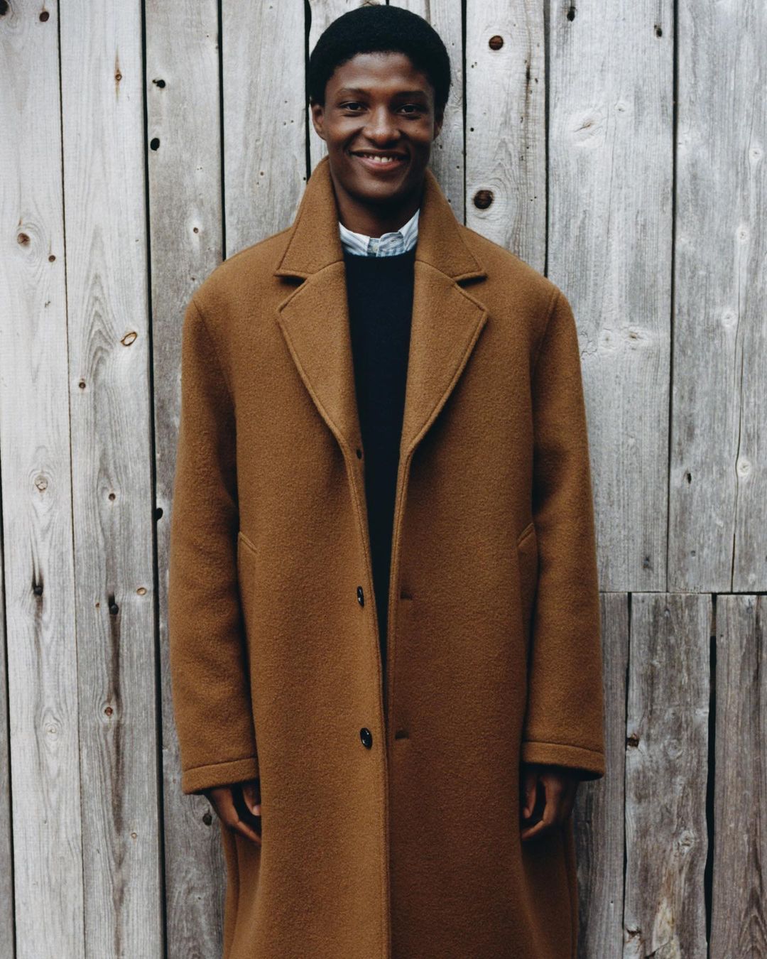 Smiling African American man in a brown wool coat stands against a rustic wooden wall for a fashion editorial portrait.