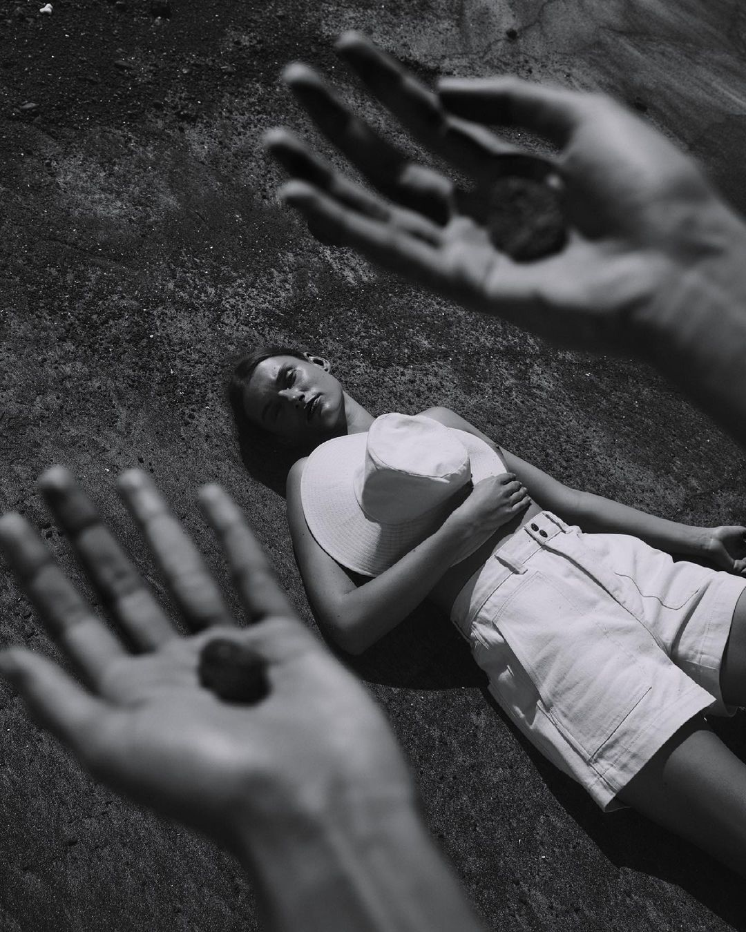 Monochrome portrait of a woman reclining on a textured surface, with foreground hands holding small stones, creating a surreal effect.