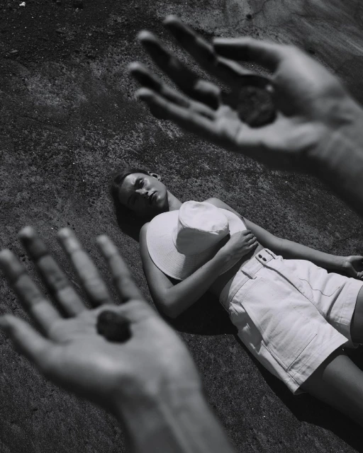 Monochrome portrait of a woman reclining on a textured surface, with foreground hands holding small stones, creating a surreal effect.