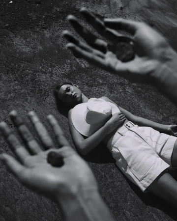 Monochrome portrait of a woman reclining on a textured surface, with foreground hands holding small stones, creating a surreal effect.