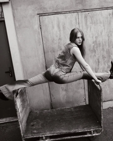 Black-and-white portrait of a young woman in jeans and a vest doing the splits on a metal cart against a concrete wall.