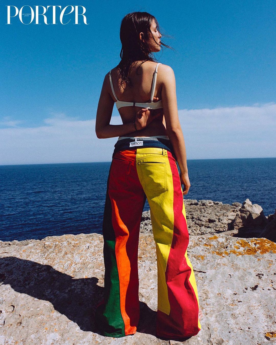 Model poses near the ocean in colorful striped pants, a white bikini top, and Porter Magazine in the upper left corner against a blue sky.