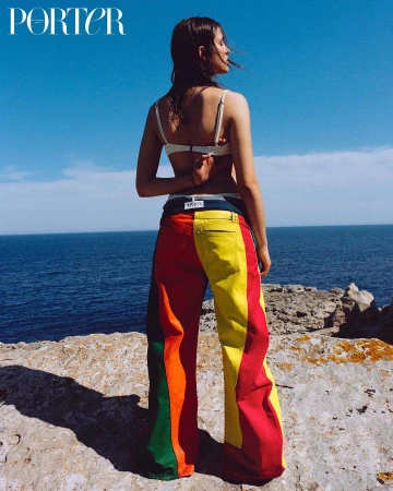 Model poses near the ocean in colorful striped pants, a white bikini top, and Porter Magazine in the upper left corner against a blue sky.