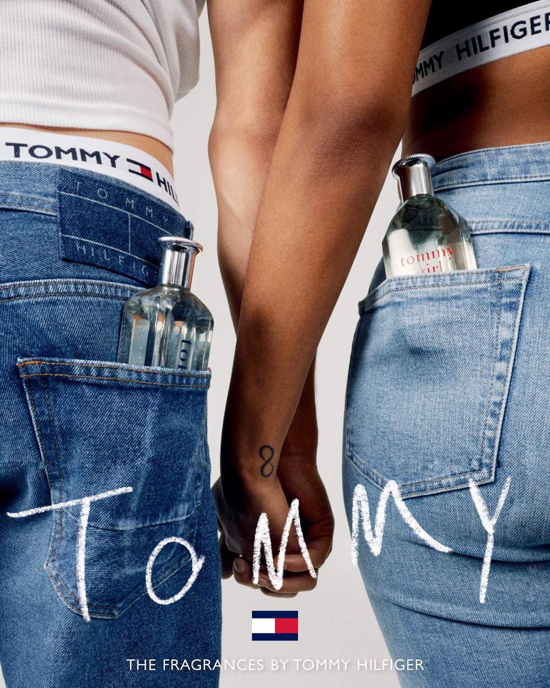 Two people model Tommy Hilfiger jeans and fragrance. Clear glass perfume bottles sit in back pockets. Close-up shot of holding hands.