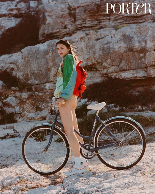 A woman poses with a bicycle in front of a rocky landscape. She wears a colorful jacket and carries a red backpack for a fashion editorial.