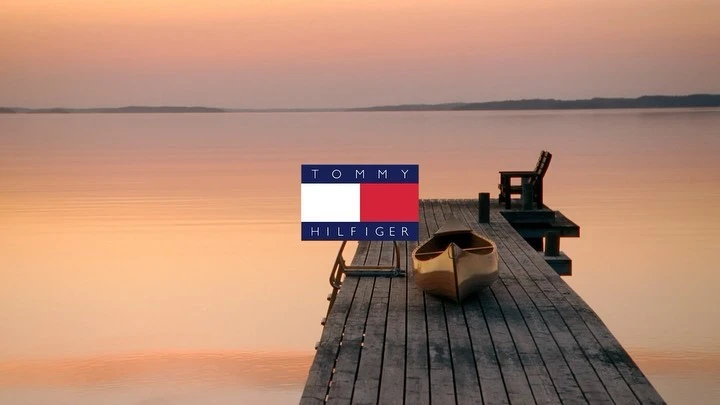 Tommy Hilfiger logo over a serene sunset lake scene with a wooden dock, boat, and chair, bathed in golden light.