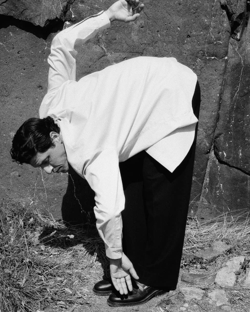 A man in a white button-down shirt bends over outdoors, reaching towards his black shoes, with a dark rock formation behind him.