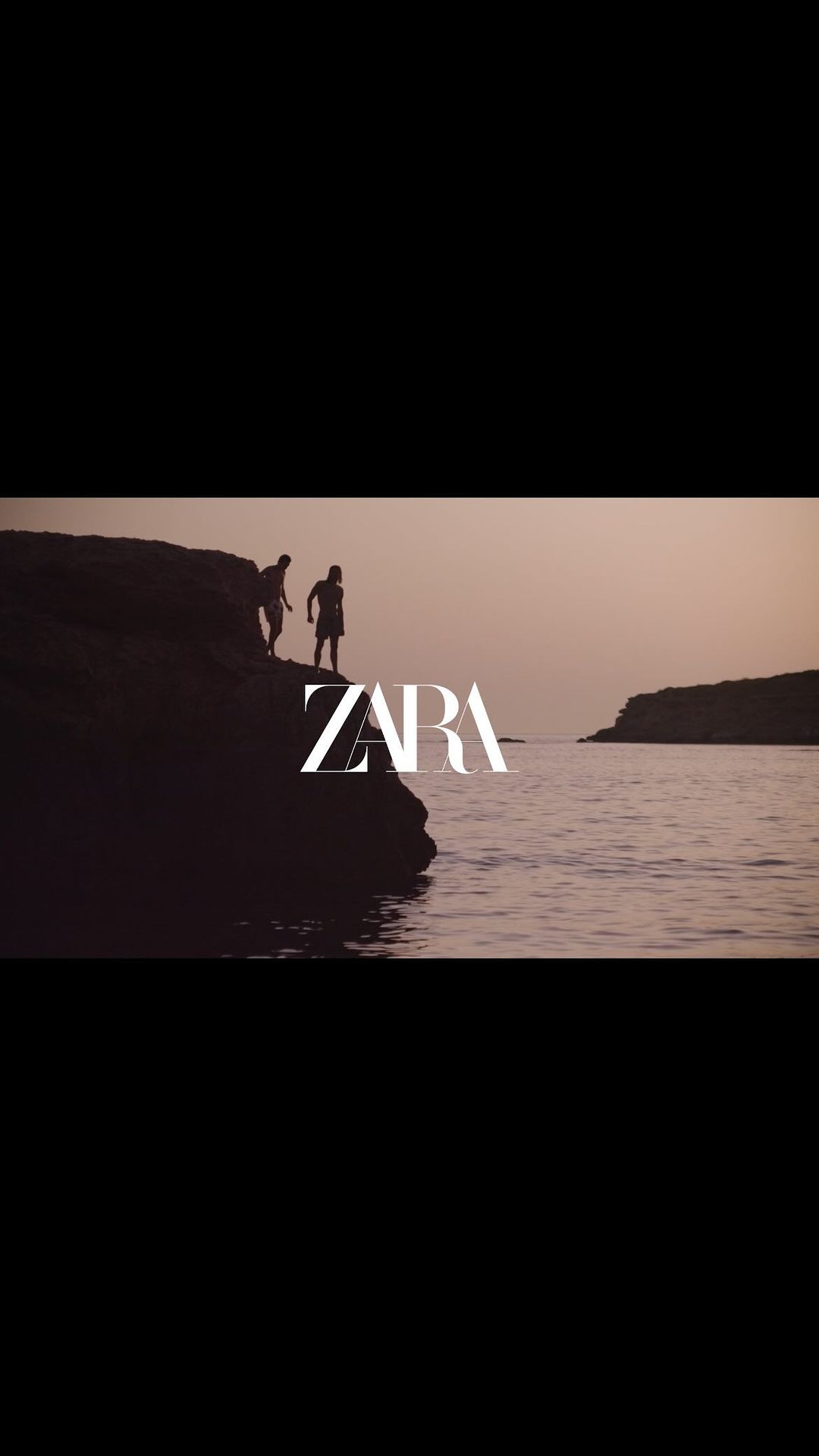 Two silhouetted figures stand on a rock overlooking a body of water, with the ZARA logo prominently displayed in the foreground.