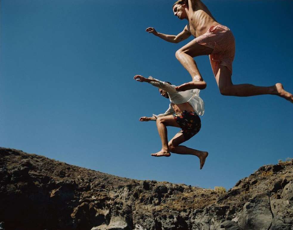 Two adult males leap mid-air with a clear blue sky background and rocky cliffs below, capturing the essence of summer freedom.
