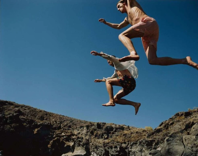 Two adult males leap mid-air with a clear blue sky background and rocky cliffs below, capturing the essence of summer freedom.