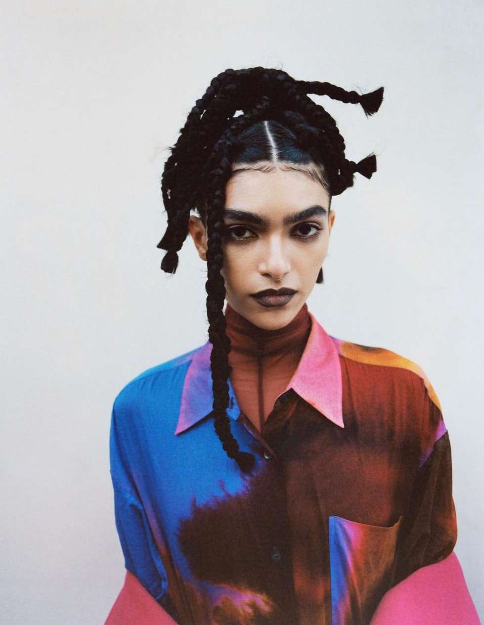 Portrait of a woman with braided hair wearing a colourful button-down shirt against a light background in a studio environment.