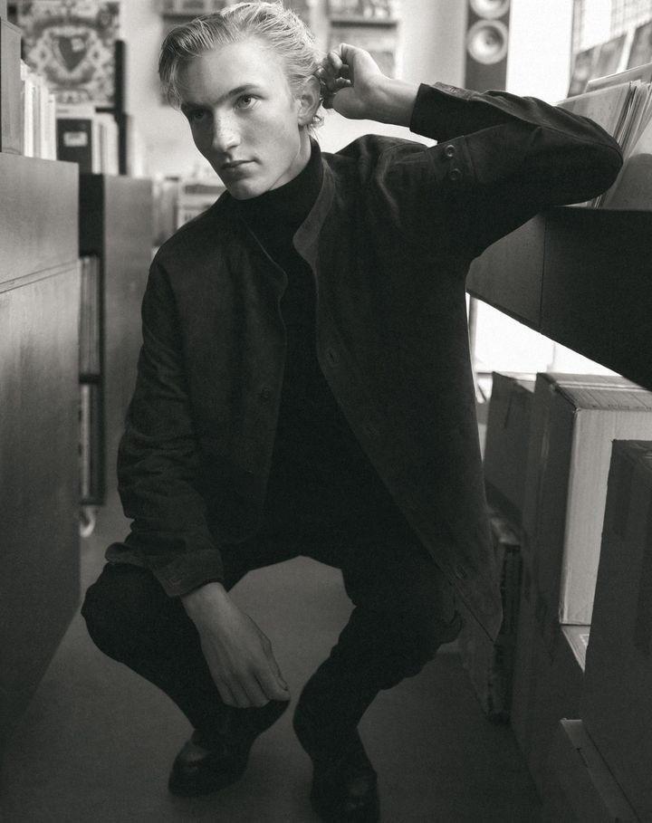 Young man with blonde hair in a dark turtleneck and jacket crouching inside a record store by shelves