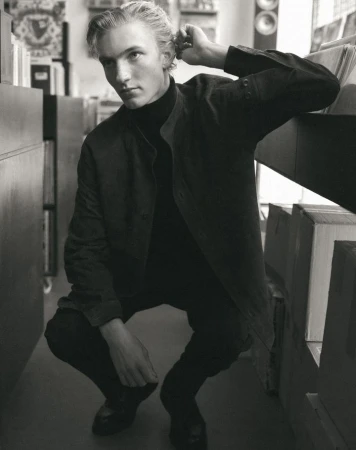 Young man with blonde hair in a dark turtleneck and jacket crouching inside a record store by shelves