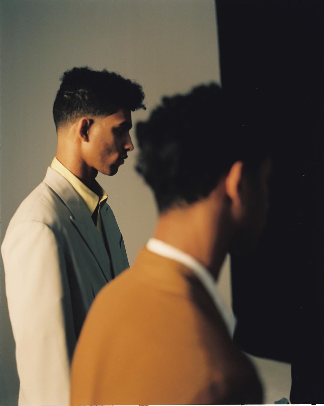 Two young men in suit jackets pose indoors against a neutral wall in a studio-style portrait with soft lighting, shot at eye-level.
