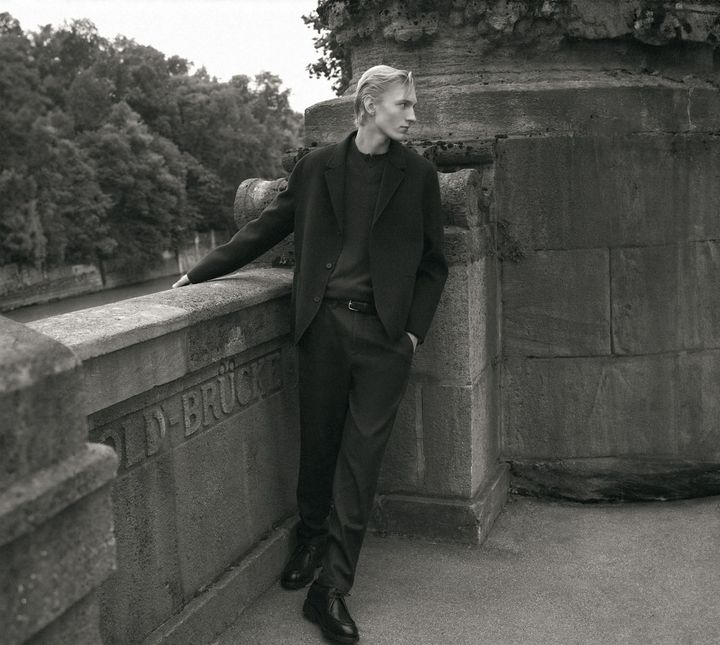 Monochrome portrait of a young man leaning against a stone bridge structure wearing a dark blazer and trousers.