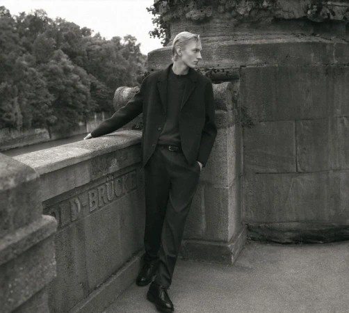 Monochrome portrait of a young man leaning against a stone bridge structure wearing a dark blazer and trousers.