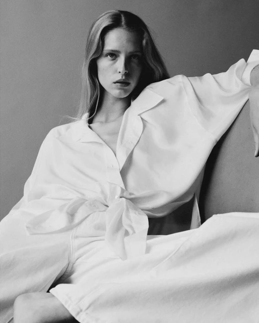 Black and white portrait of a young adult woman with long hair, wearing a white shirt and pants in a minimalist studio setting.