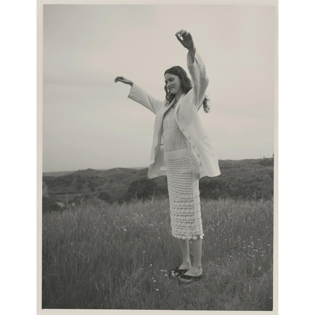 Woman in linen suit stands with arms raised in grassy field against hilly background, wearing a knitted skirt and flat shoes.