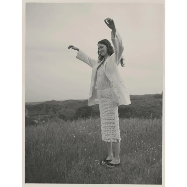 Woman in linen suit stands with arms raised in grassy field against hilly background, wearing a knitted skirt and flat shoes.