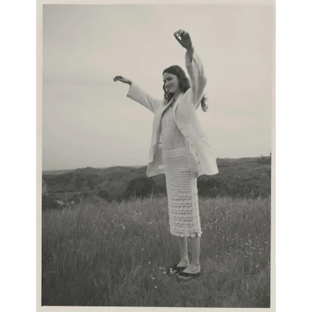 Woman in linen suit stands with arms raised in grassy field against hilly background, wearing a knitted skirt and flat shoes.