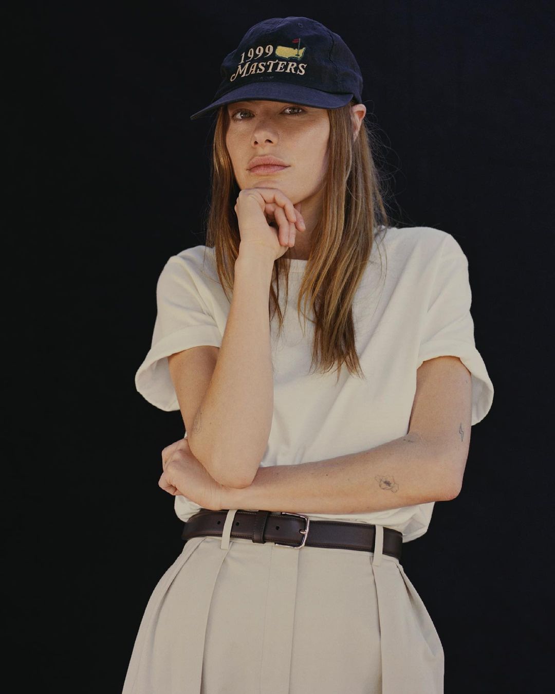 Woman in a navy baseball cap and white T-shirt poses in front of a black background. She wears khaki pants and a brown leather belt.