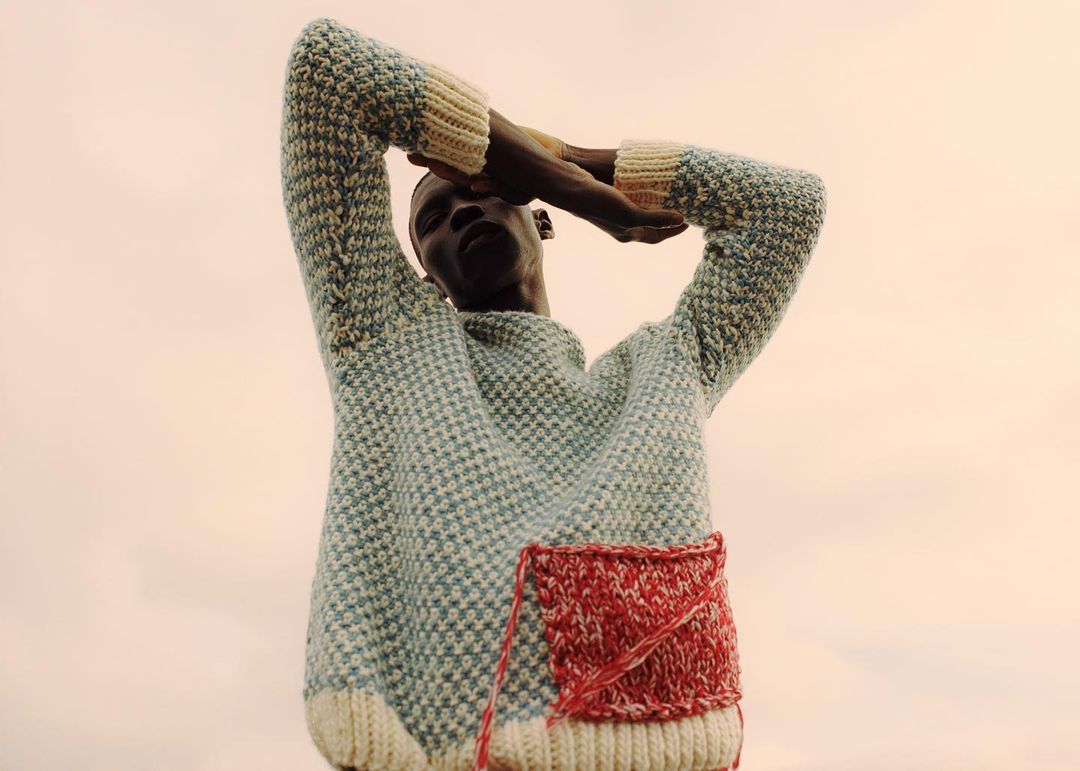 Portrait of a dark-skinned male model wearing a knitted sweater with his arms raised against a neutral background.
