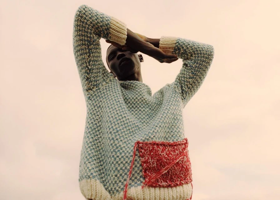 Portrait of a dark-skinned male model wearing a knitted sweater with his arms raised against a neutral background.