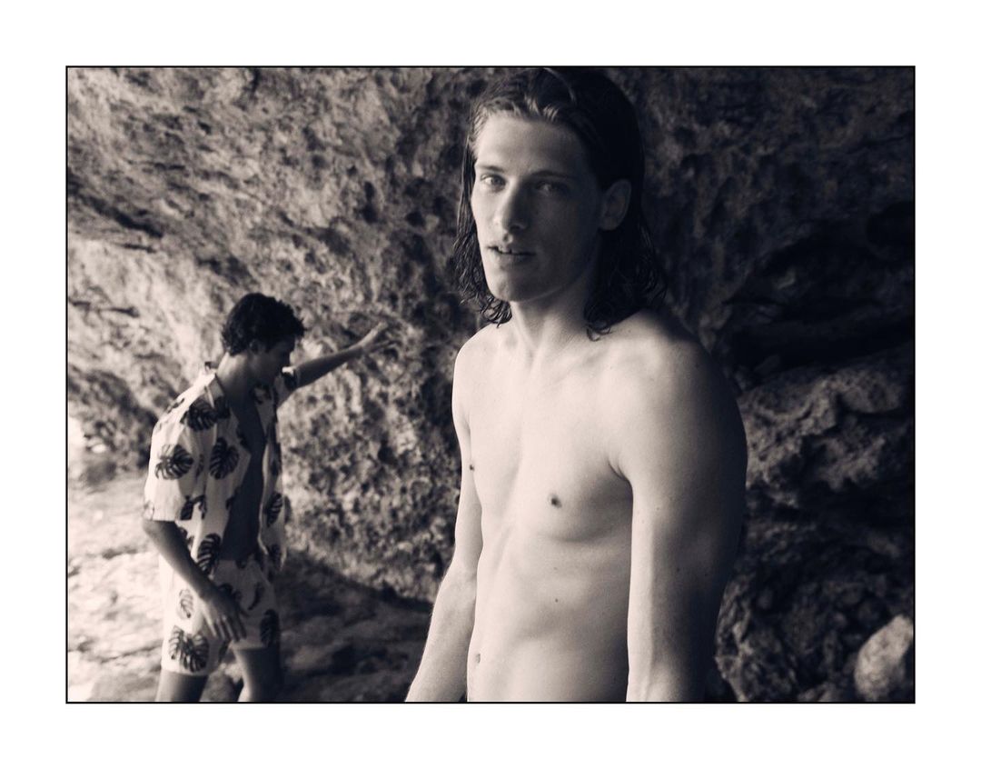 Monochromatic shot of shirtless man standing near a man in tropical shirt and shorts with rock formation in background, outdoors.