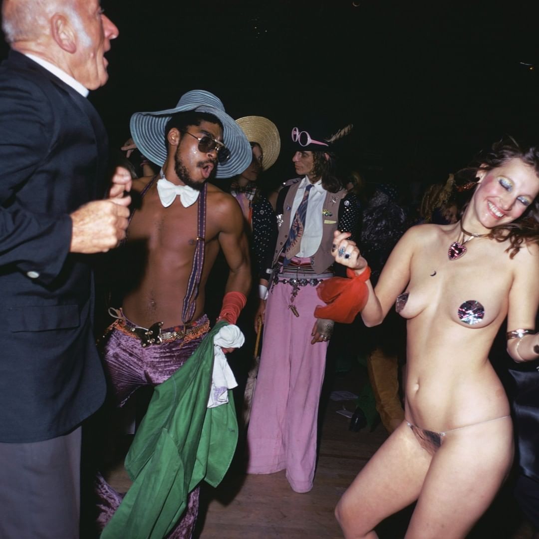 A crowd of people dances at a lively disco party with colorful outfits, retro fashion, and energetic movement in a dark indoor setting.