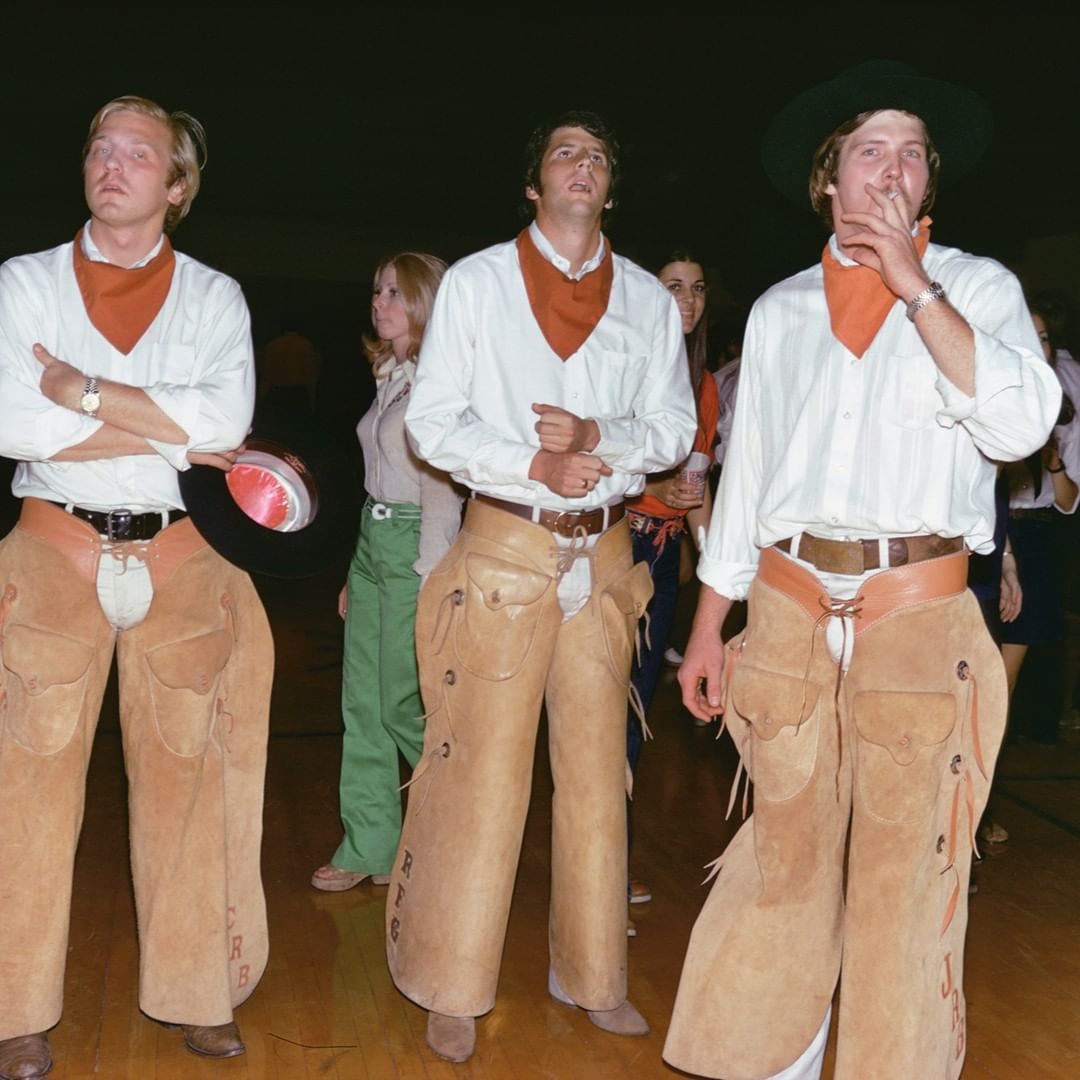 Three young men wearing cowboy chaps, white shirts, and orange neckerchiefs stand indoors in a group, with other people in the background.
