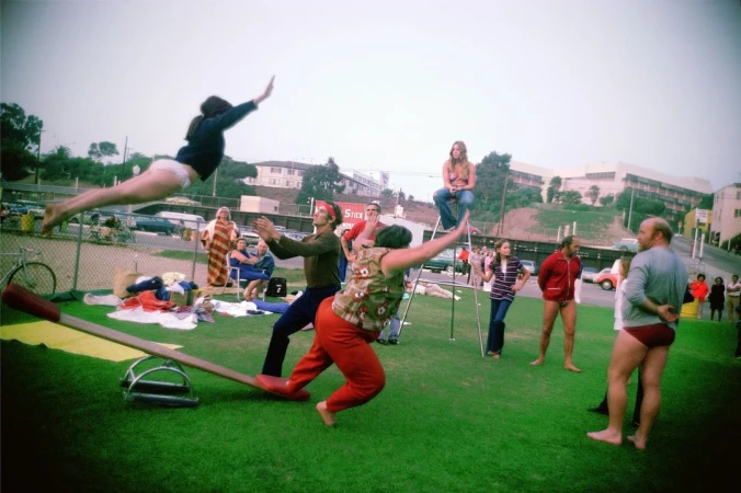 Outdoor gathering with a person jumping over a seesaw, surrounded by other adults on green grass with buildings in the background.