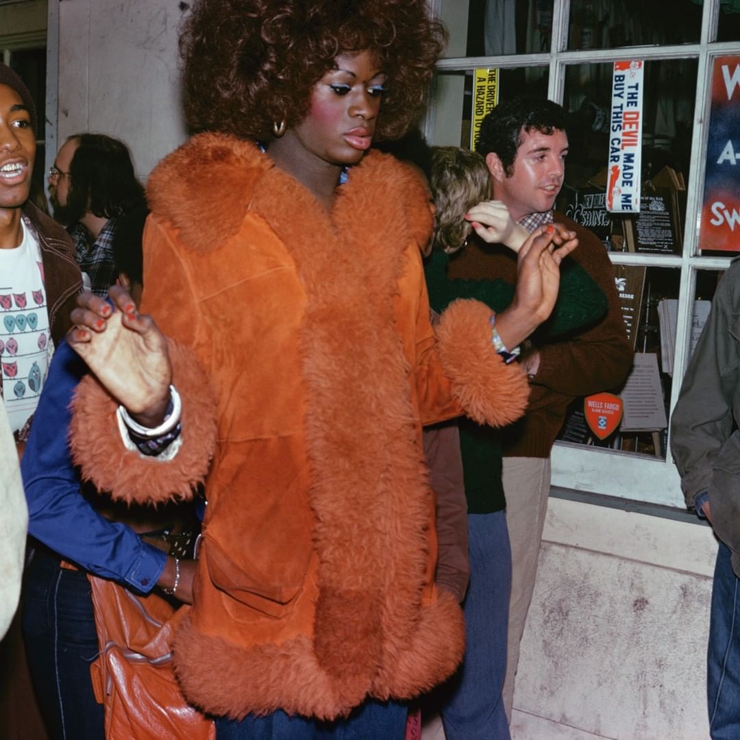 Portrait of a non-binary person in an orange fur-trimmed coat, standing among a group of people outside near a building at night.