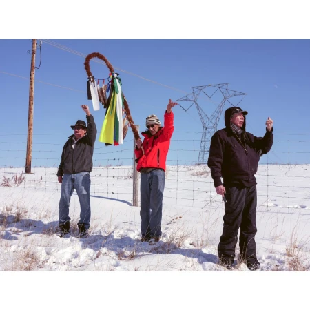 Three adults in outerwear stand in a snowy field beneath power lines, one holding a ribboned object, under a blue sky in a wide shot.