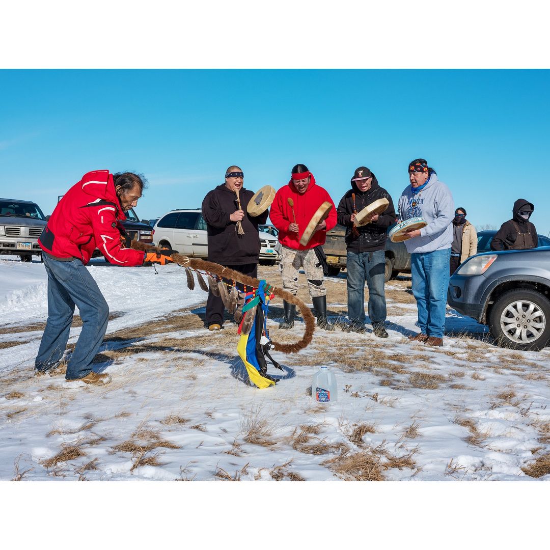 A group of Indigenous adults plays drums on a snow-covered field, under a clear sky. The photo captures a traditional outdoor gathering.