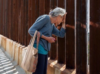 Elderly woman peers through steel bars of border fence, wearing blue sweater and carrying a tote bag in an outdoor setting.