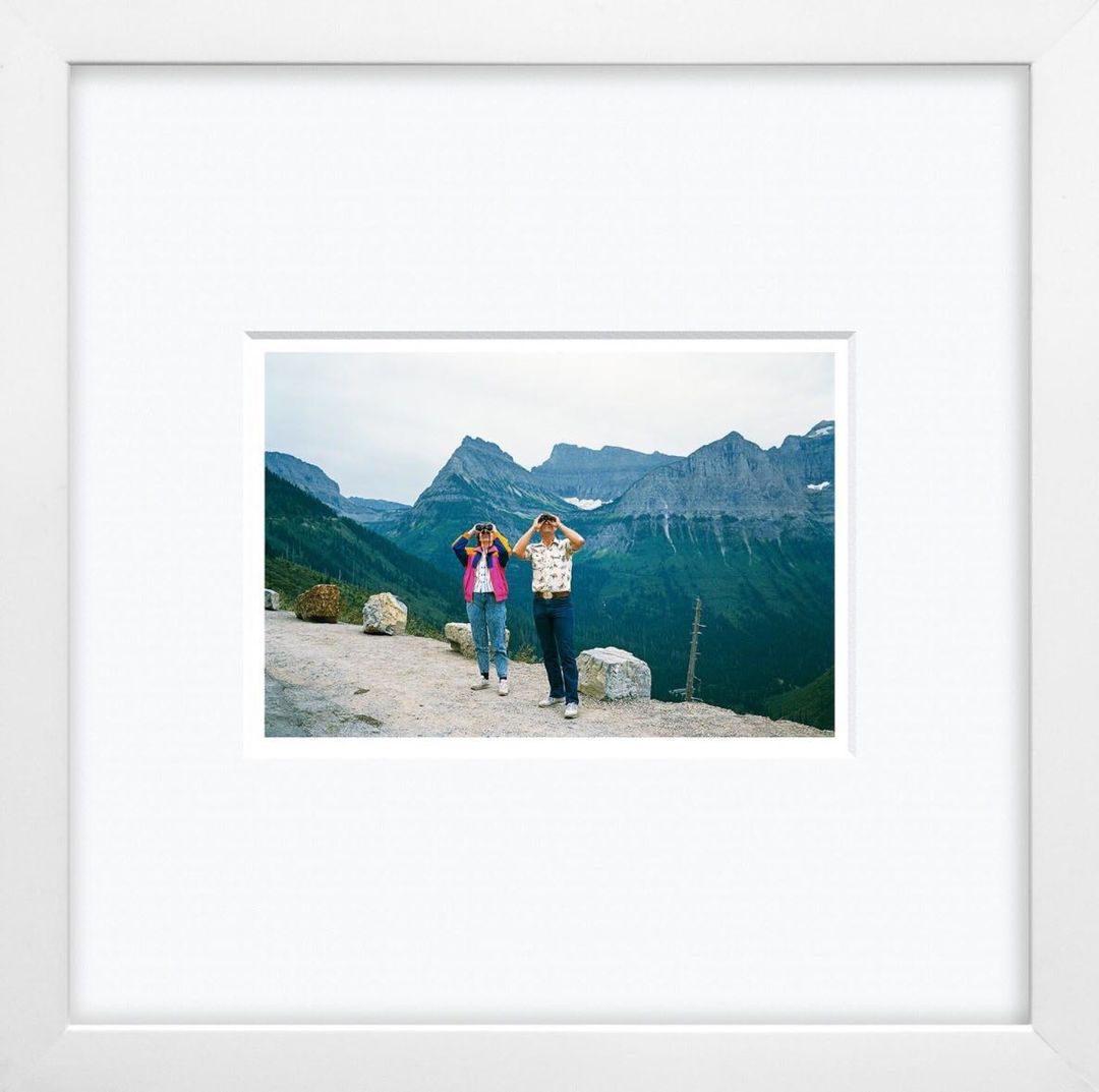 Two people pose against a mountain backdrop, with their hands raised to their heads, framed by a white border and a white frame.