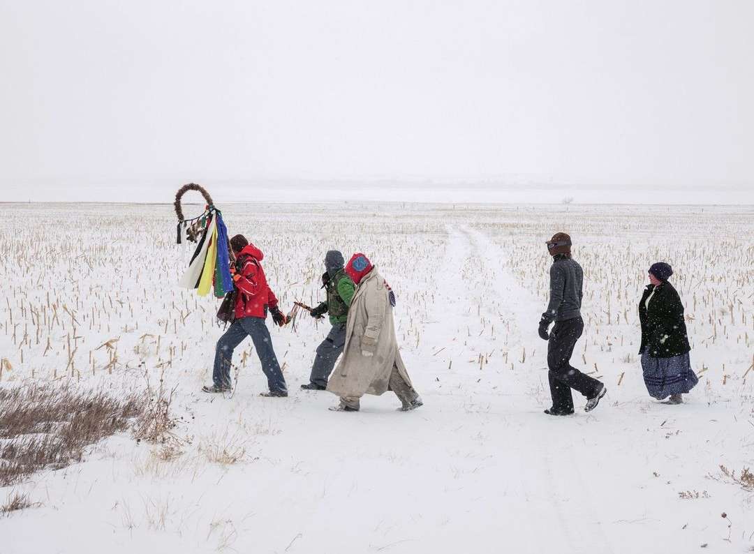 A group of five people walk through a snow-covered field, with a sparse winter landscape under an overcast sky, in a wide outdoor shot.