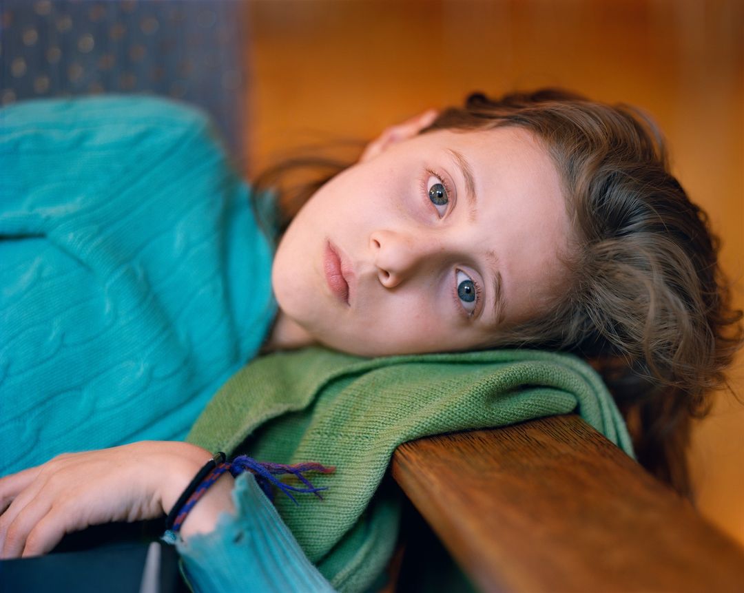 A young girl with blue eyes and brown hair rests her head on her arms, wearing a teal sweater in soft, natural light.