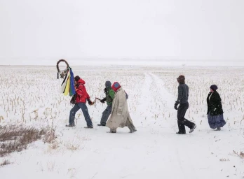 Five people walking across a snow-covered field under an overcast sky carrying a decorated staff.
