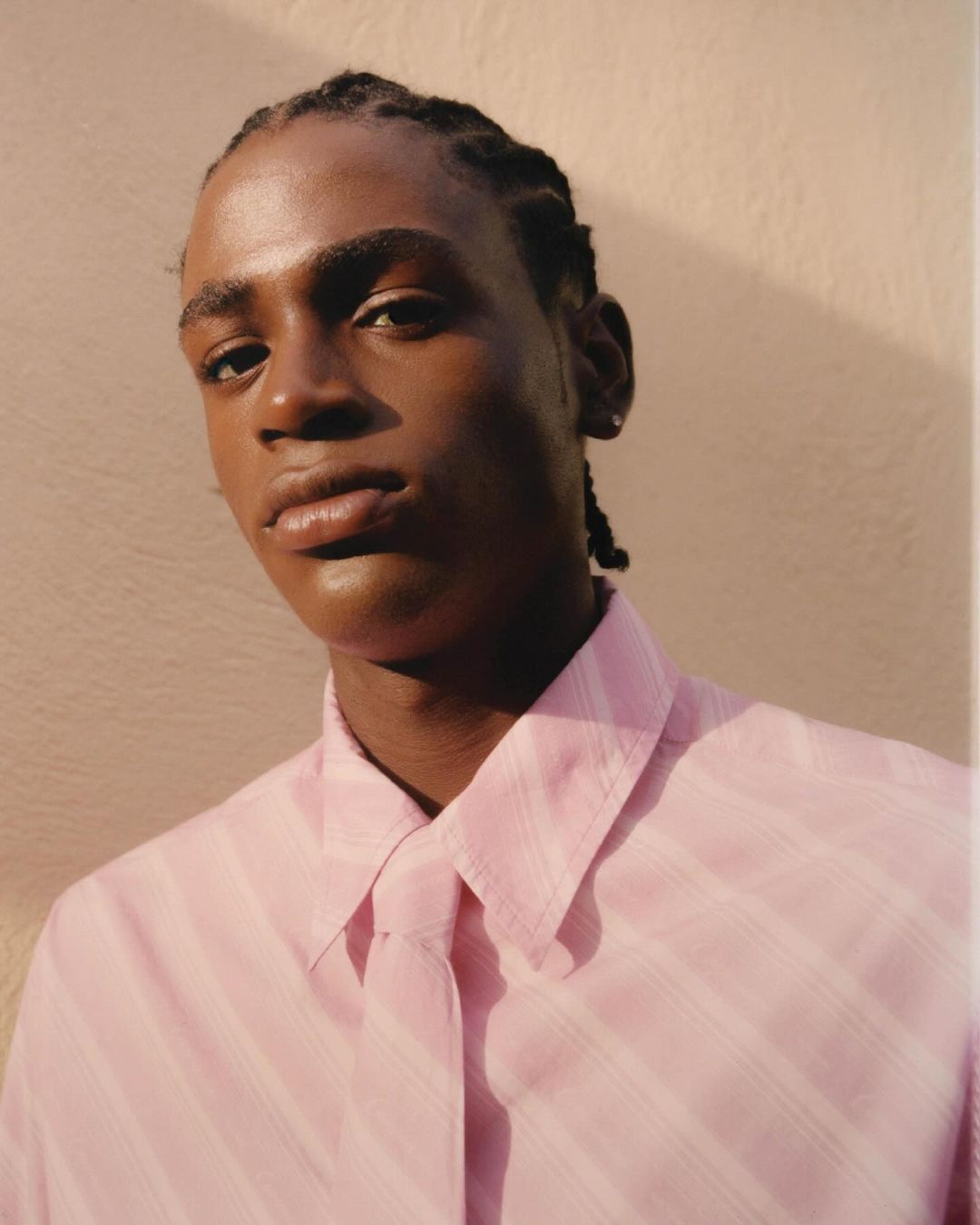Portrait of a young adult male with braided hair, wearing a light pink striped button-down shirt against a neutral wall, in soft light.
