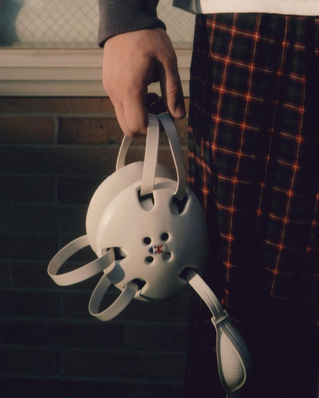 Close-up of a person holding a white wrestling headgear with red accents, set against a backdrop of brick and plaid patterns.