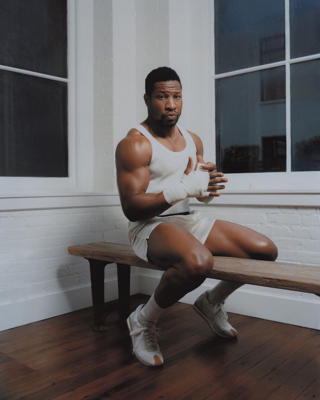 Portrait of a muscular adult male sitting on a bench, wrapping his hands, indoors with natural window lighting.
