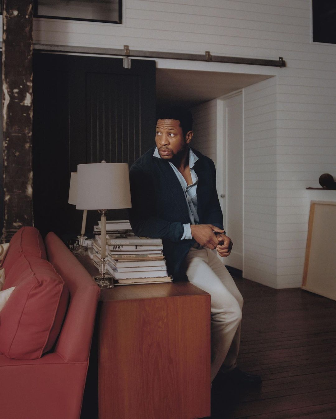 Adult male in a blue blazer and cream pants is sitting on the corner of a wooden desk in a modern interior space, looking off-camera.