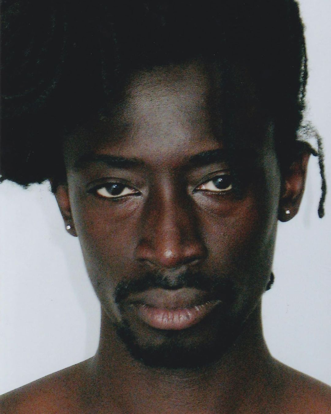 Close-up portrait of a Black man with dark hair and a mustache, looking directly at the camera against a plain background.