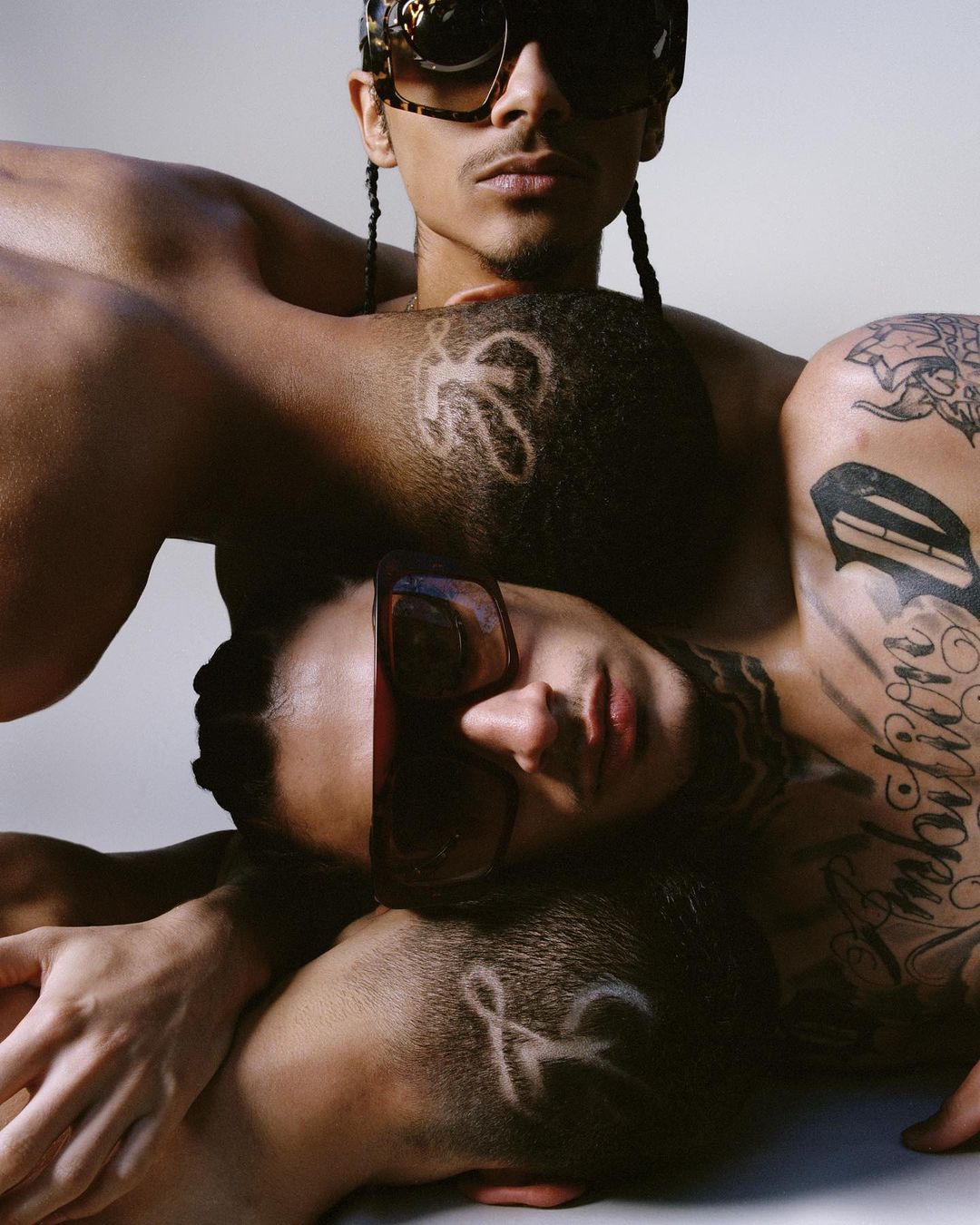 Artistic studio portrait of three shirtless male models with sunglasses in a close-up composition against a neutral backdrop.