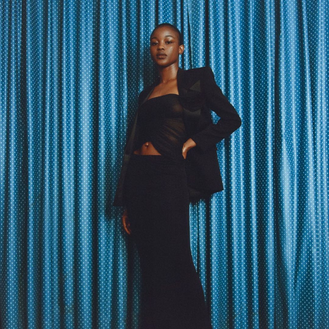 Portrait of a Black female fashion model in a strapless top and blazer, posing against a dotted blue curtain backdrop in a photo studio.