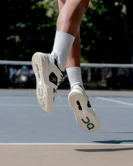 Low angle view of person in athletic shoes jumping above blue tennis court with green trees in background on a sunny day.